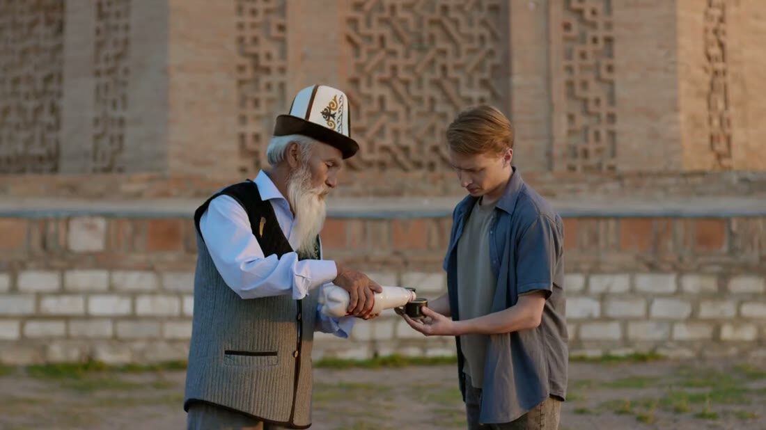 Live-action still — Bakai pours kymyz for Alex by the ornamented wall
